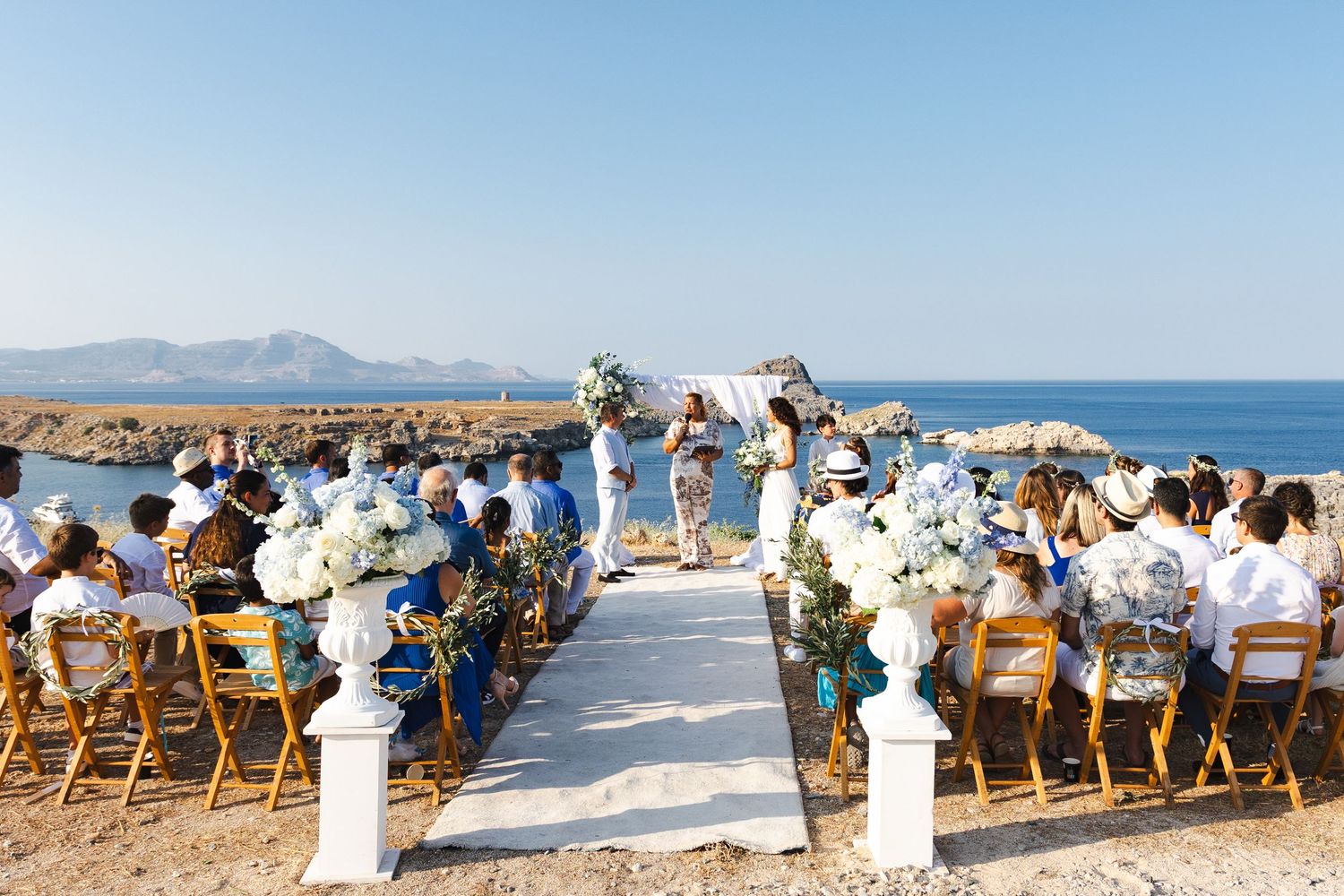 A scenic beach wedding ceremony setup with wooden chairs and ocean views along a rocky Mediterranean coastline.