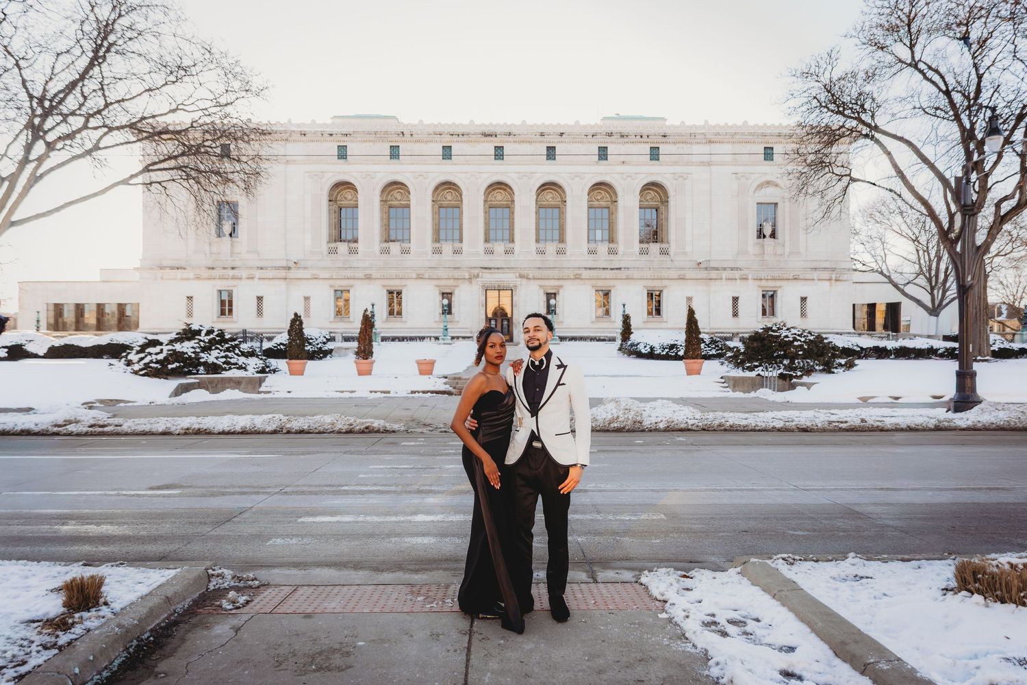 A couple stands in front of a grand historic building with snow on the ground during winter.