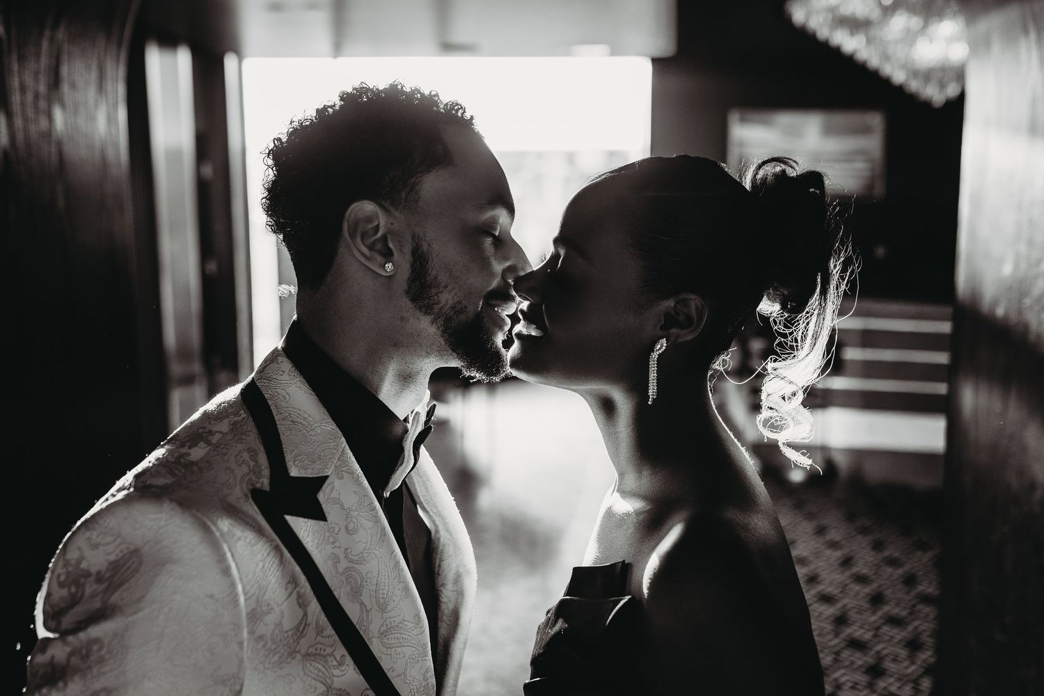 A romantic black and white photograph captures two silhouettes sharing a tender kiss in a dimly lit hallway.