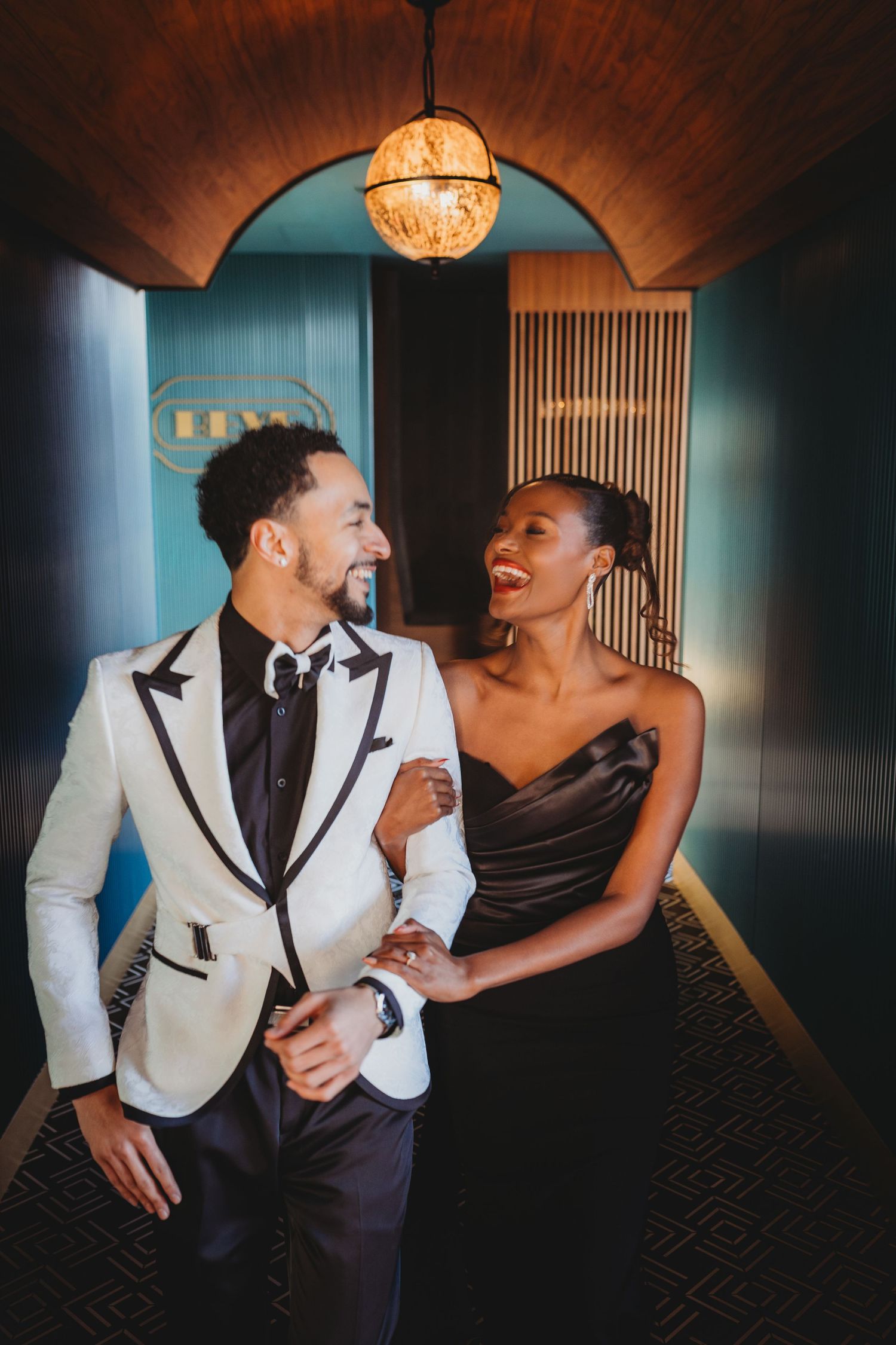 A couple in formal attire shares intimate moments while posing against a moody blue-lit wall in an elegant setting.
