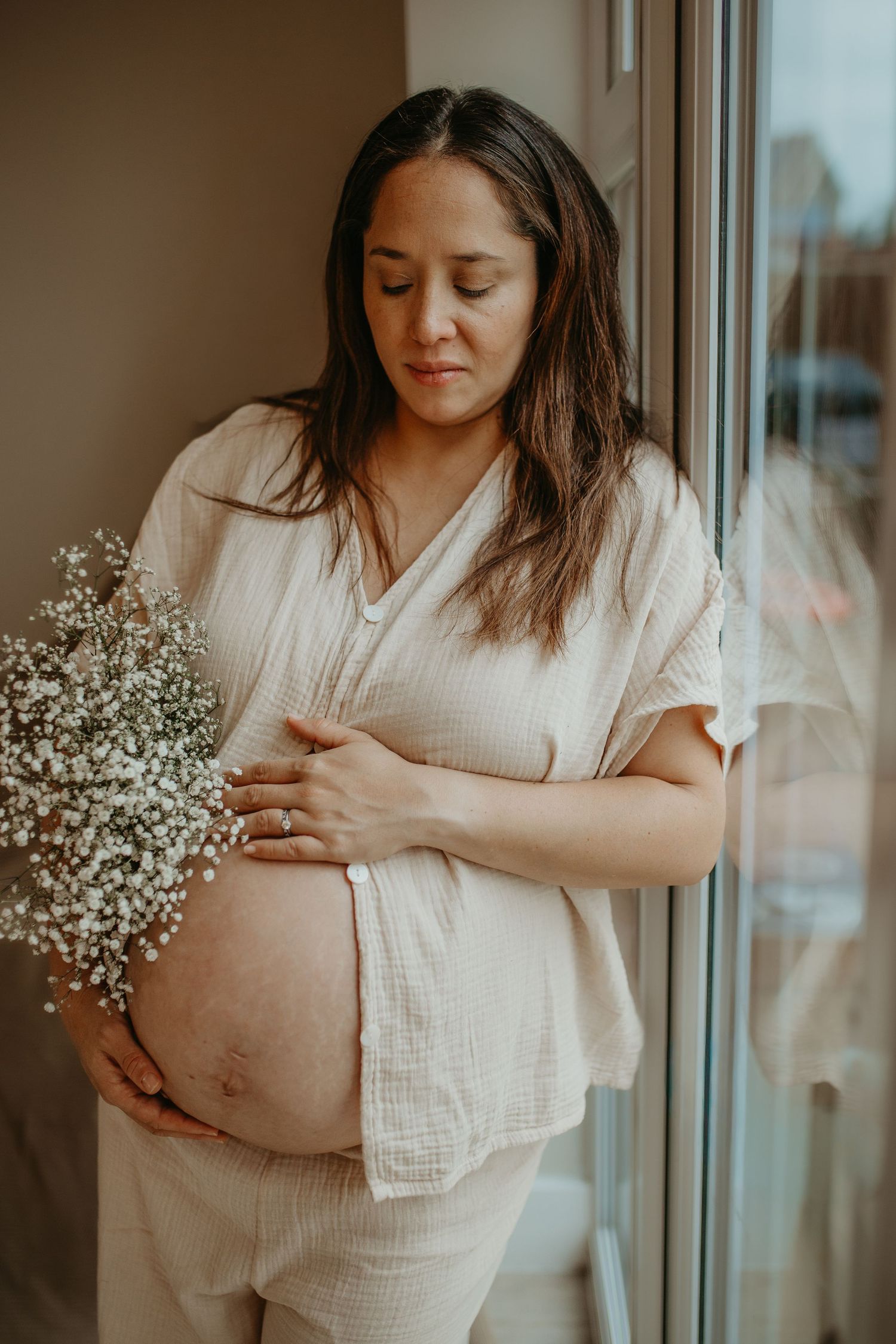 Expectant woman in white linen clothing holds baby's breath flowers while standing by window in warm lighting.