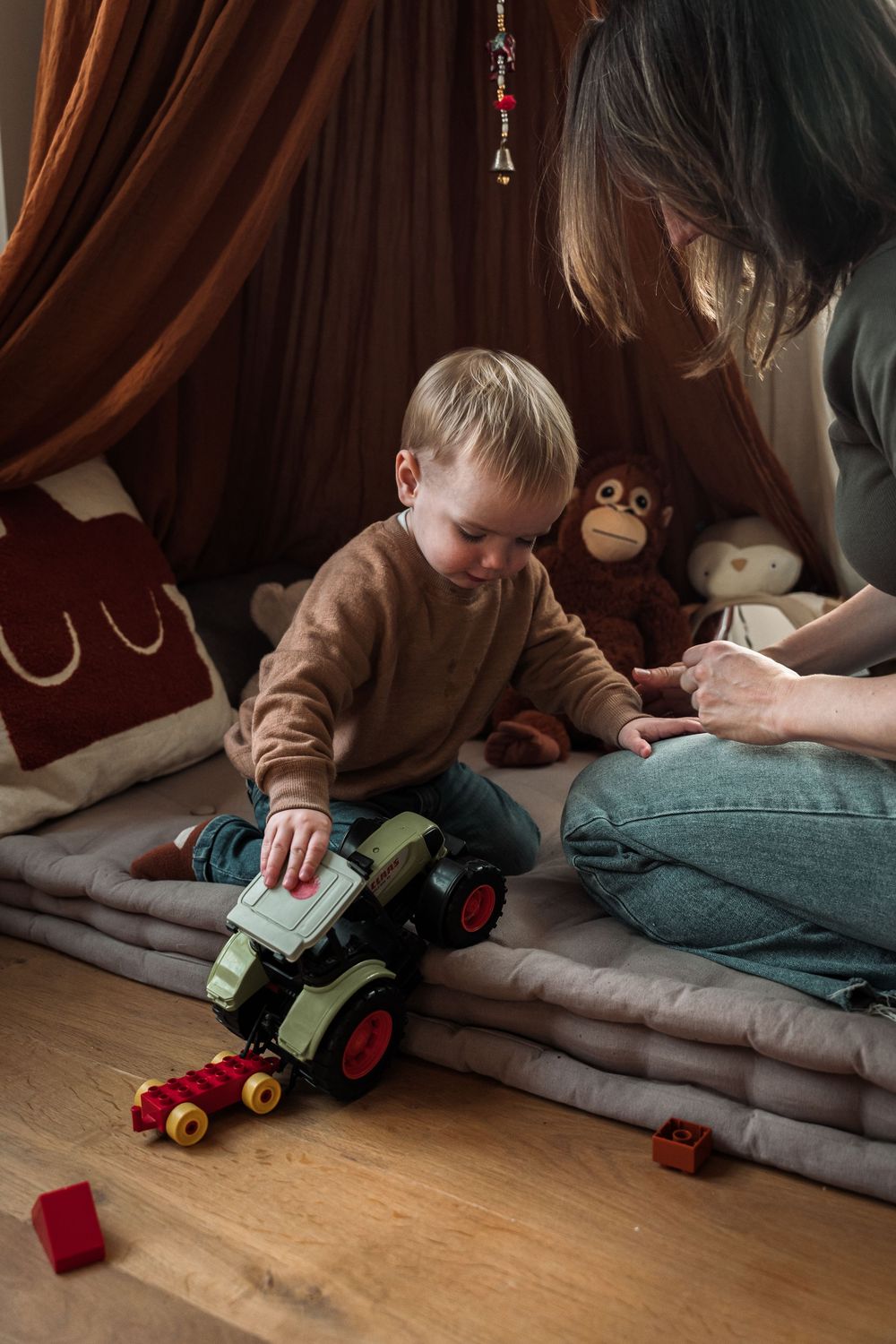 A child plays with a green toy tractor against a backdrop of brown curtains and wooden flooring.