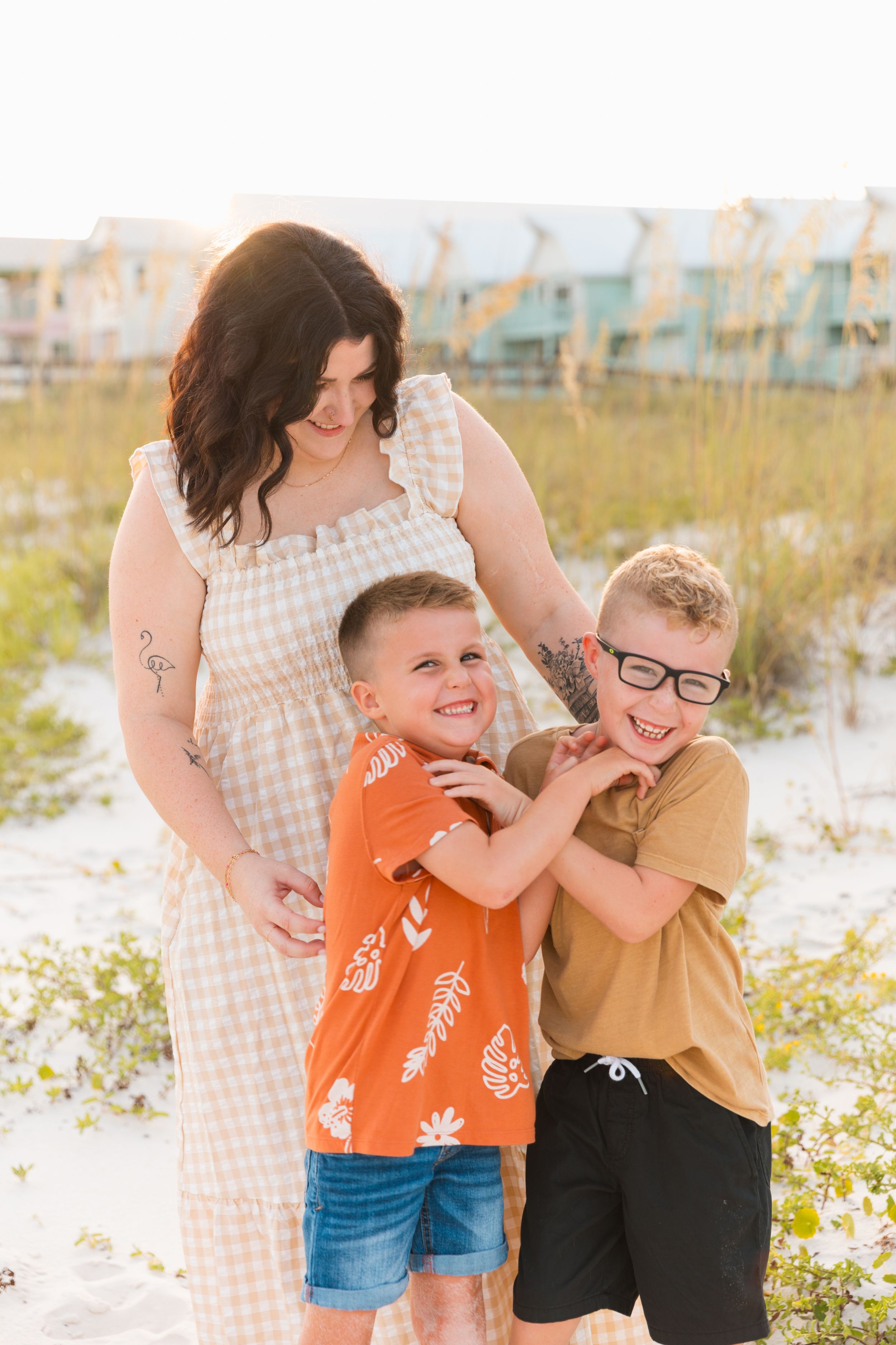 A family plays and laughs together on a sunny beach with sand dunes and coastal buildings in the background.