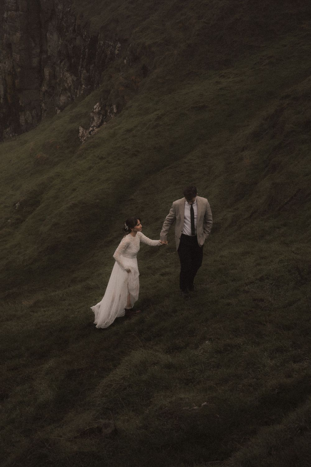A couple in wedding attire walks together on a grassy hillside slope at dusk.