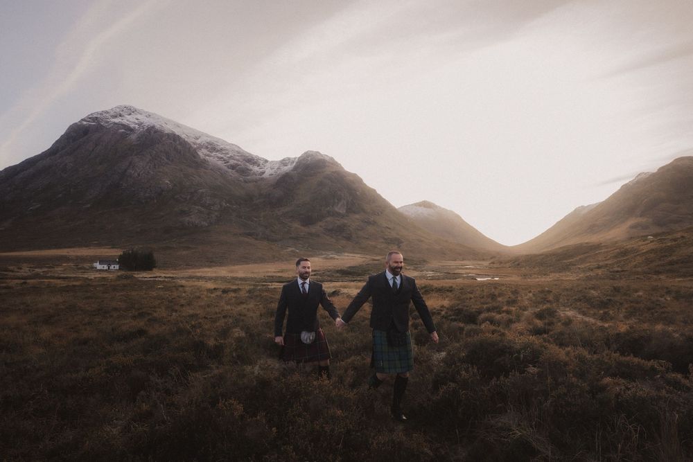 A romantic wedding couple stands silhouetted against a moody, mountainous Scottish Highland landscape at sunset.
