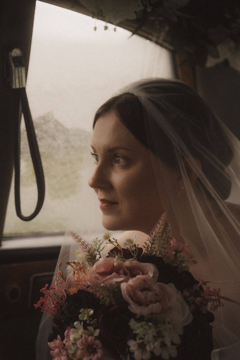 A person in a white dress sits in a car with a pink and white floral bouquet.