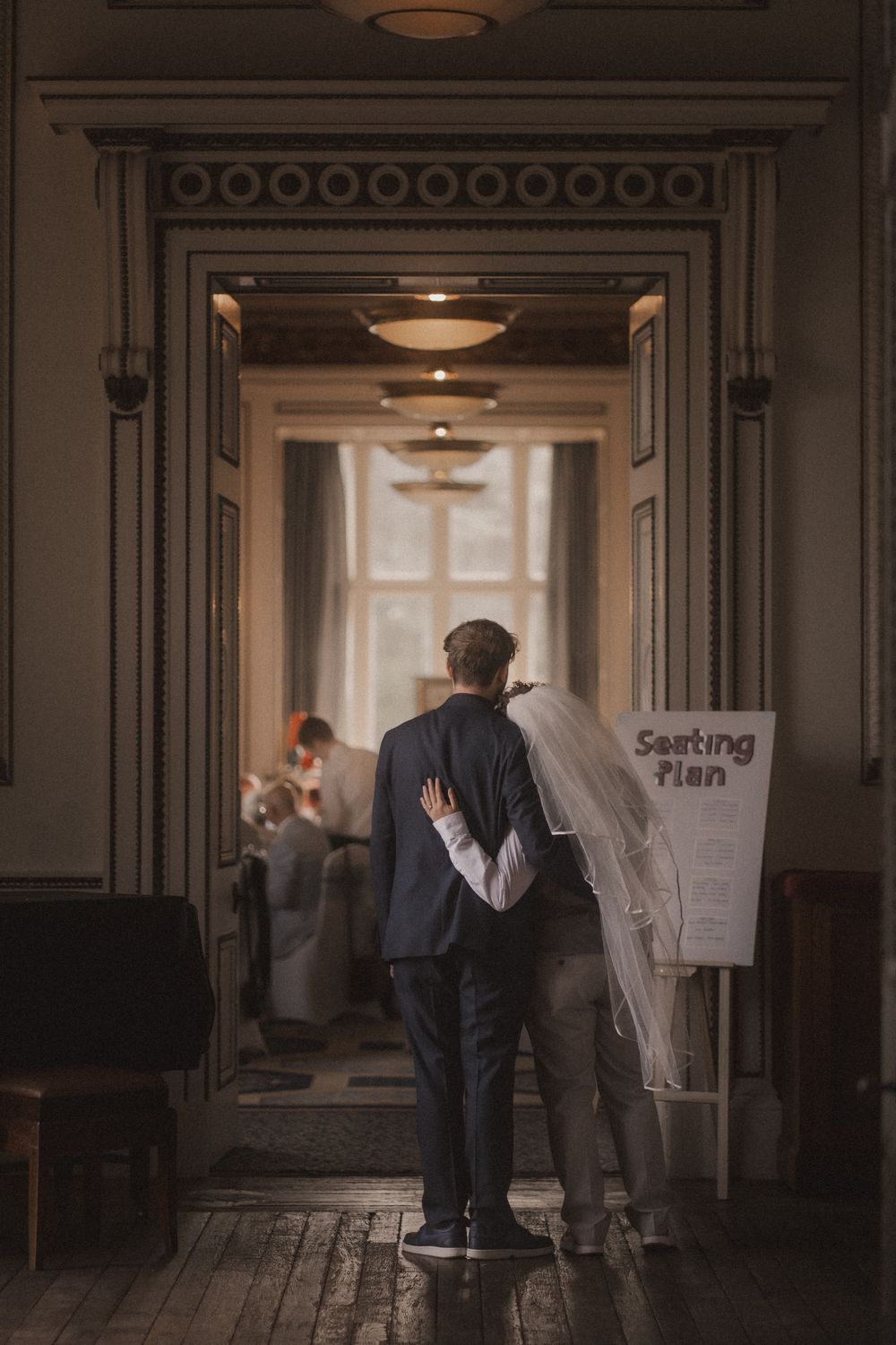 Two people stand in a dimly lit hallway looking at papers posted on a wall in an institutional setting.