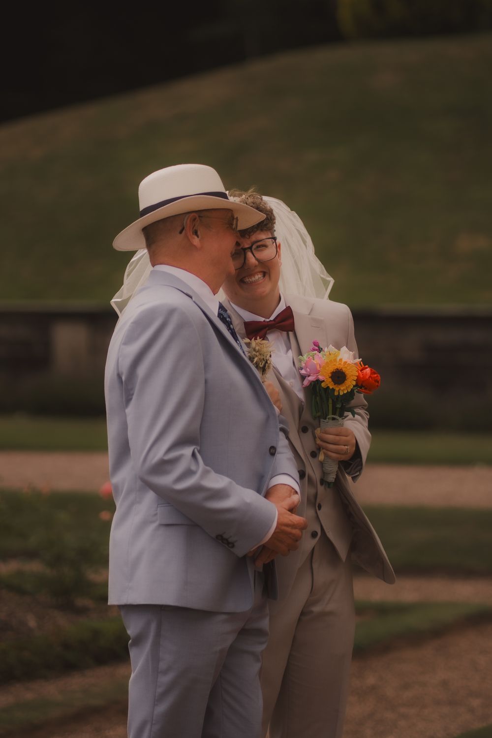 A couple in wedding attire shares a tender embrace while holding a bouquet of sunflowers and orange flowers outdoors.