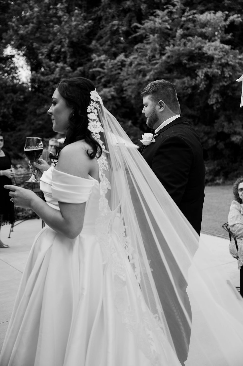 A black and white wedding photo showing a bride in an off-shoulder gown and veil holding a wine glass.