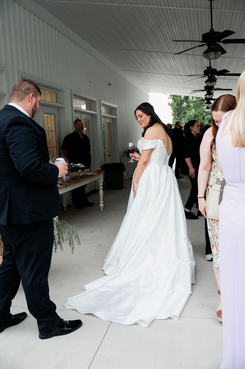 A bride in a flowing white off-shoulder gown stands in a covered porch area during her wedding reception.