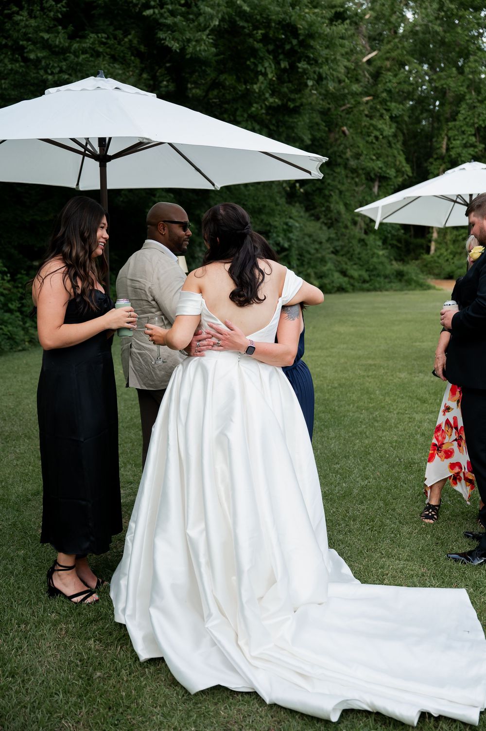 Wedding guests gather under white umbrellas on a lawn, surrounding a bride in a flowing white gown.