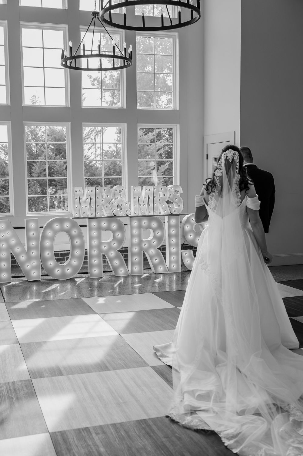 Black and white photo of wedding couple dancing in elegant venue with chandeliers.