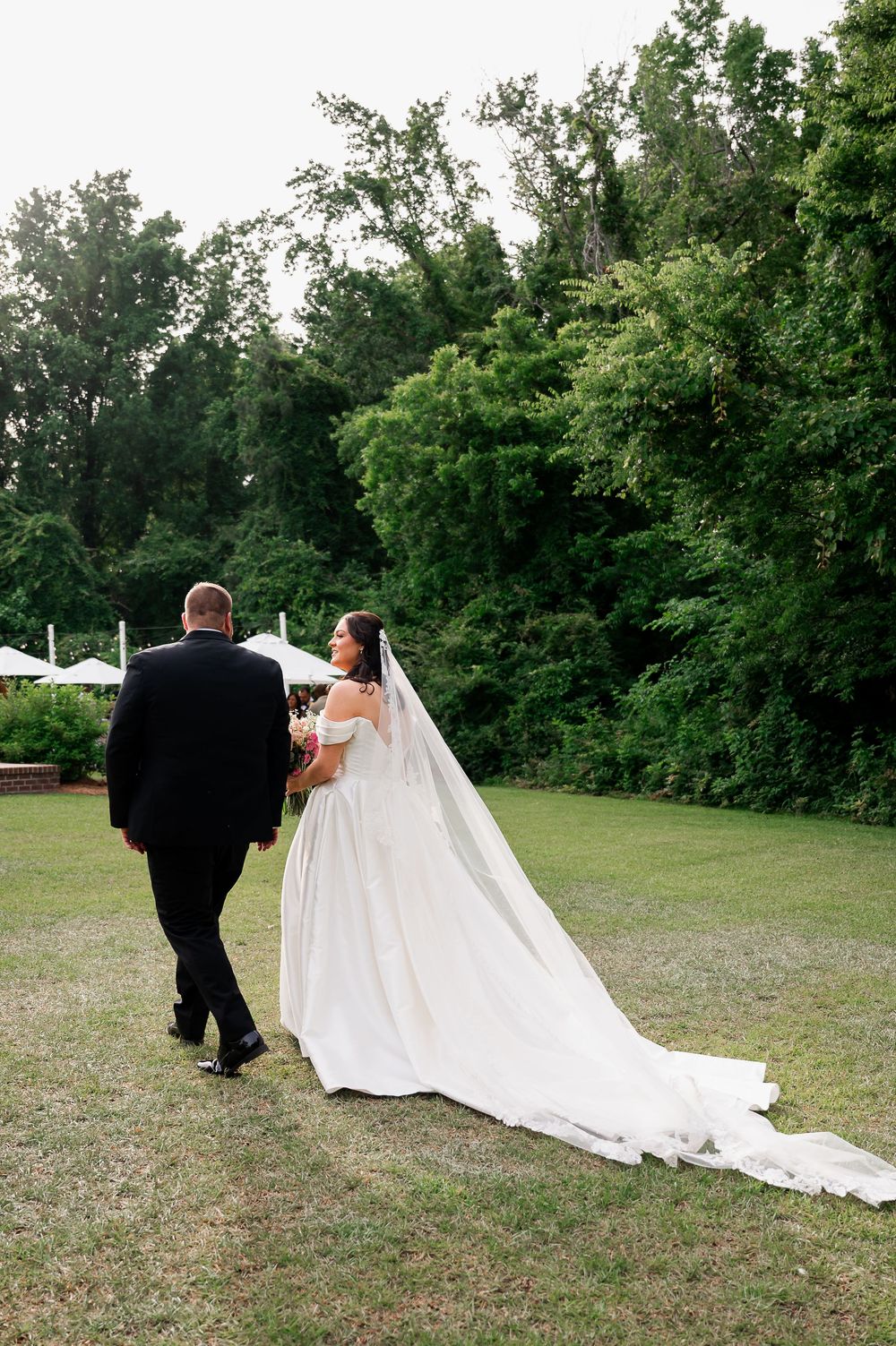 Wedding couple walking across grassy field with flowing white dress train.