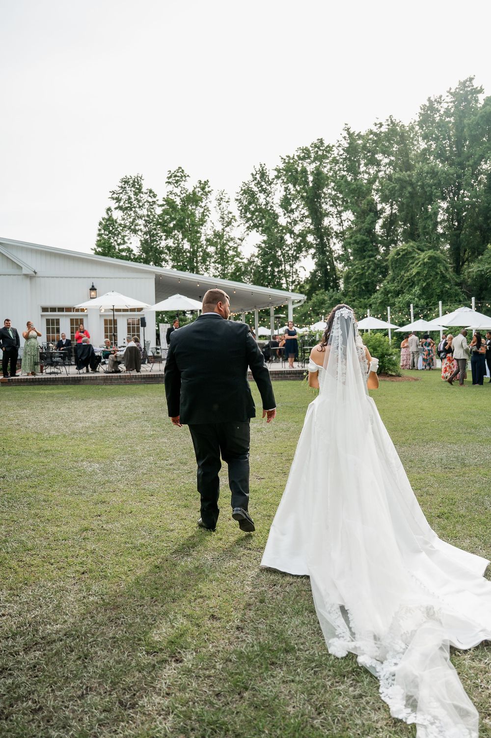 Wedding couple walking across outdoor venue grounds with flowing white dress train.