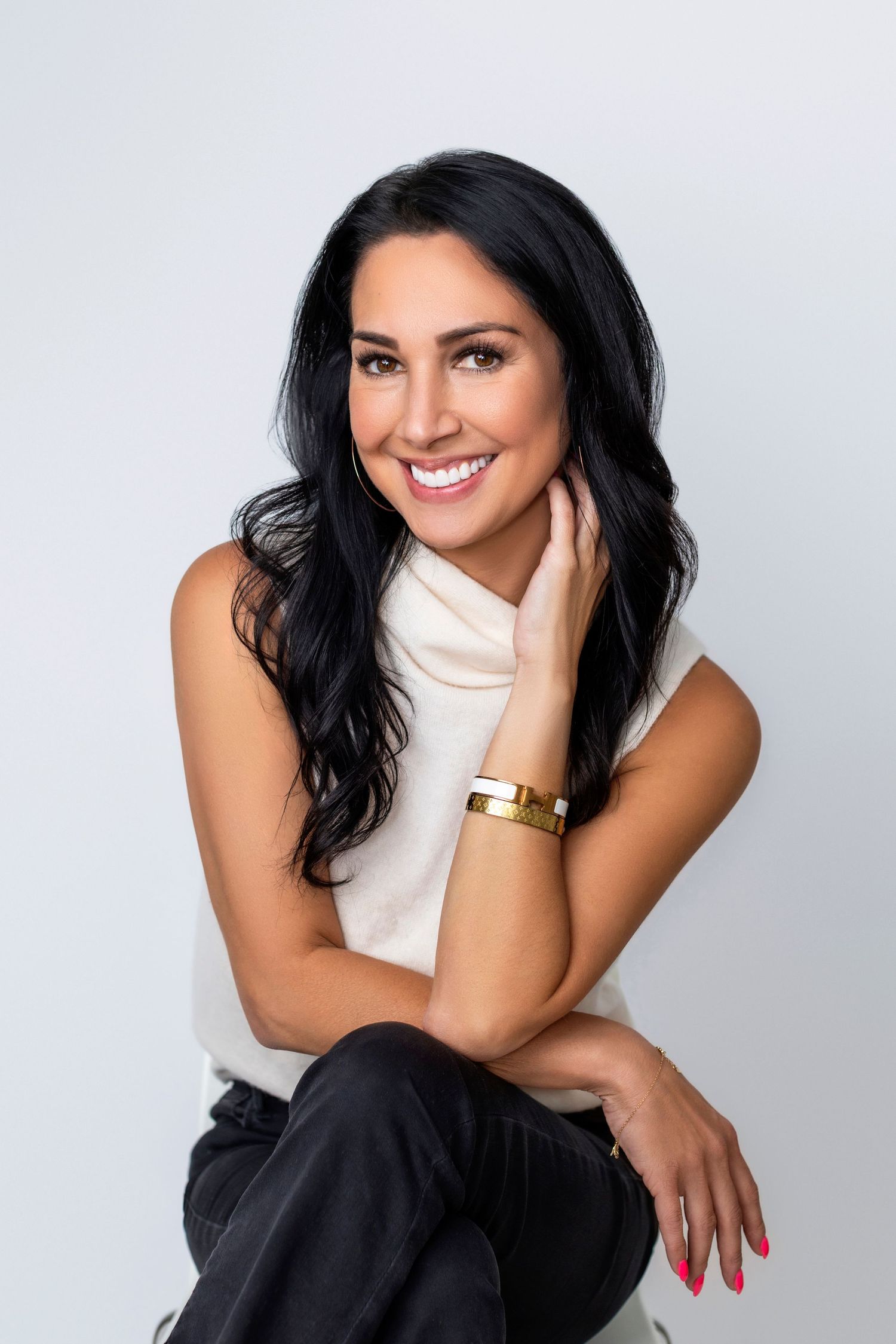 Professional studio portrait in white turtleneck and dark pants with natural makeup and dark hair against light background.
