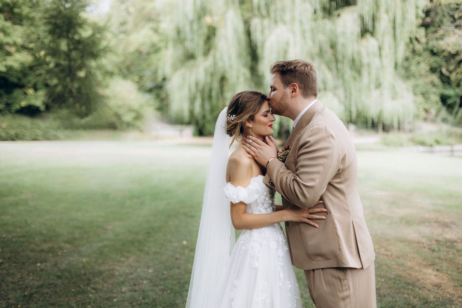 A romantic wedding couple embraces under a weeping willow tree in a lush garden setting.