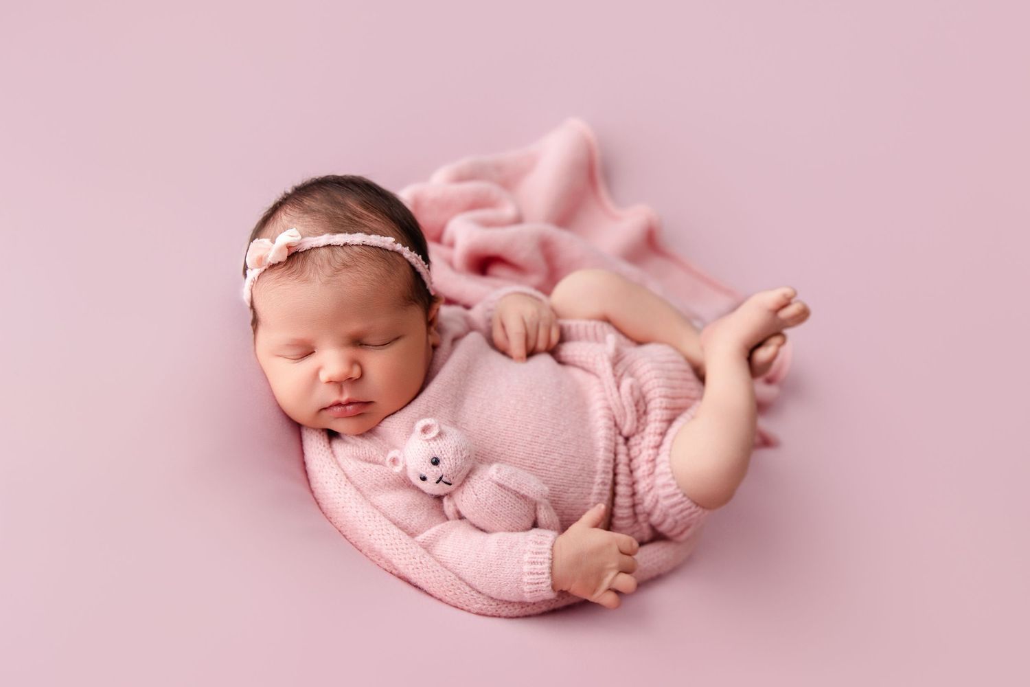 Sleeping newborn baby in pink swaddle blanket curled up in peaceful pose against soft pink backdrop.