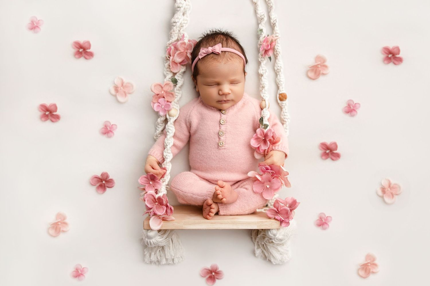 A newborn baby sleeping peacefully on pink fabric surrounded by delicate pink floral petals in a dreamy studio setting.