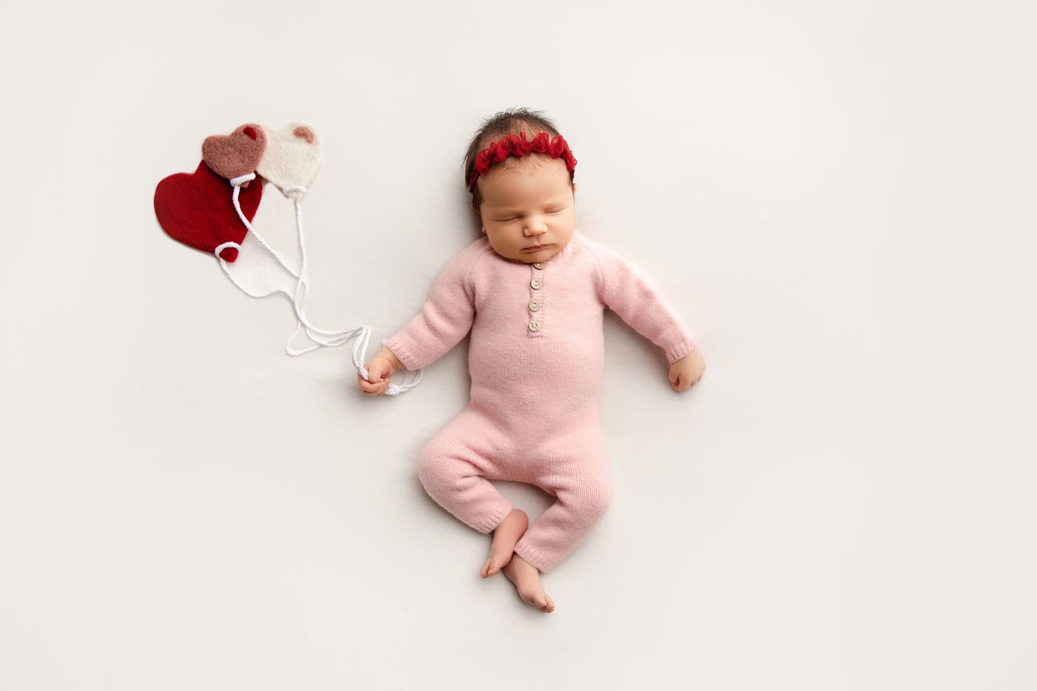 Sleeping baby in pink onesie lying on white backdrop with heart-shaped red balloons.