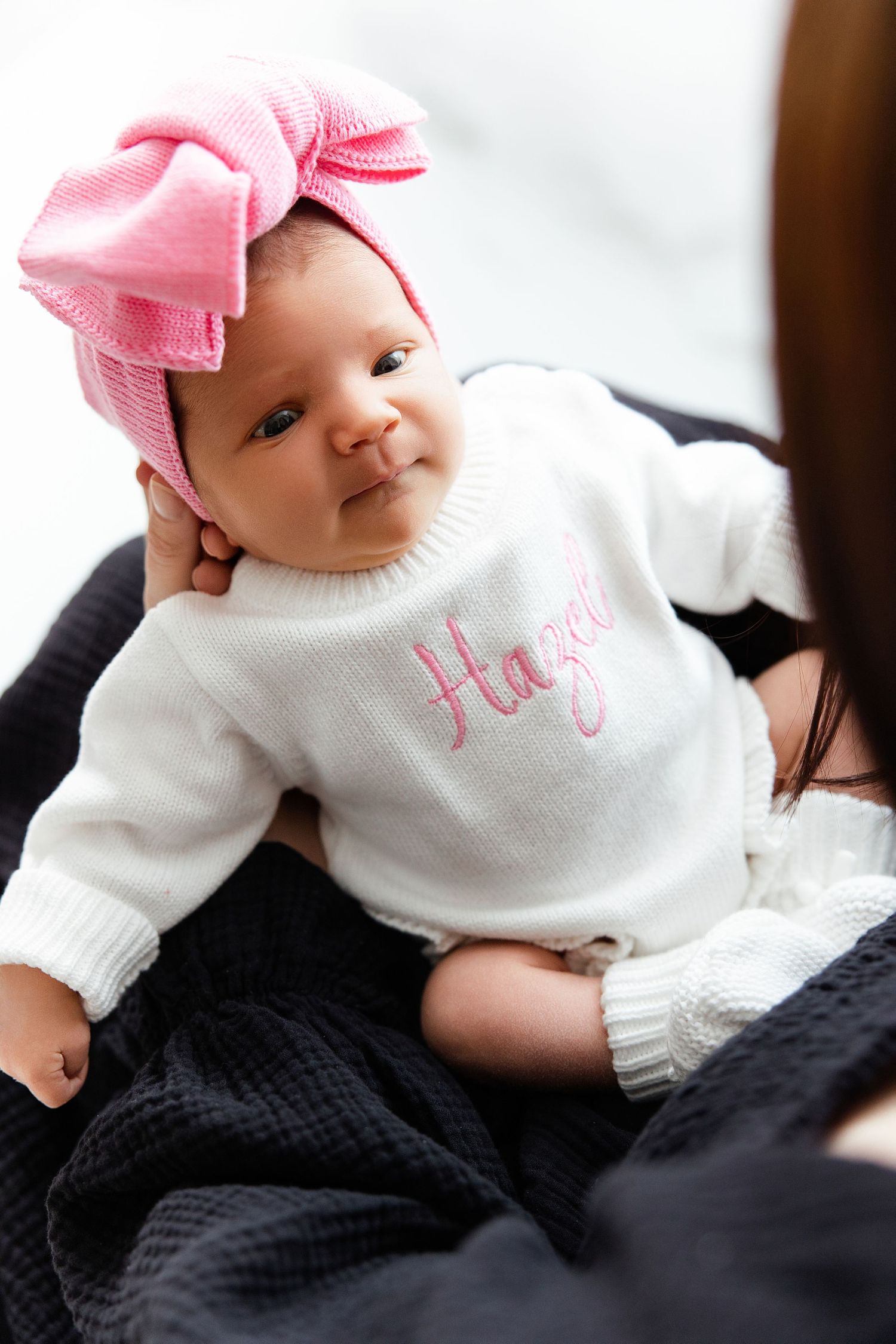 Baby wearing white sweater and pink bow headband resting on dark textured blanket.
