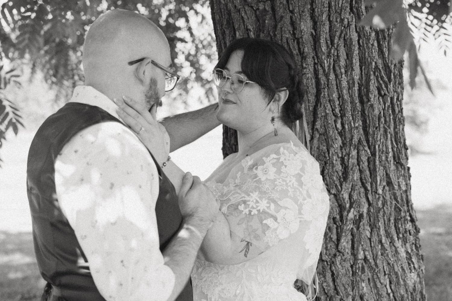 A romantic black and white wedding photo of a couple sharing an intimate moment beside a large tree trunk.
