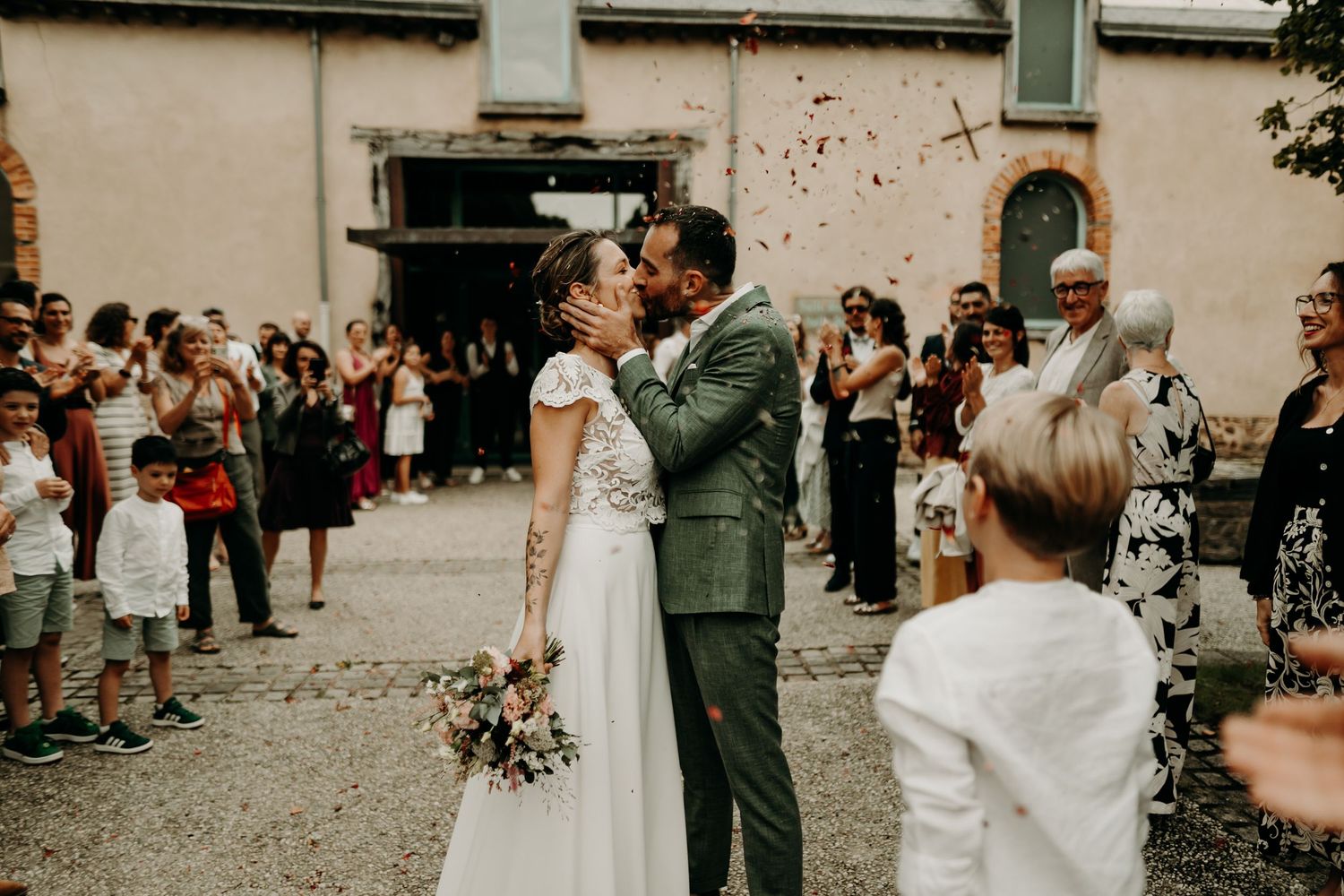 Newly married couple exits grand church building while guests throw confetti in warm sunset lighting.