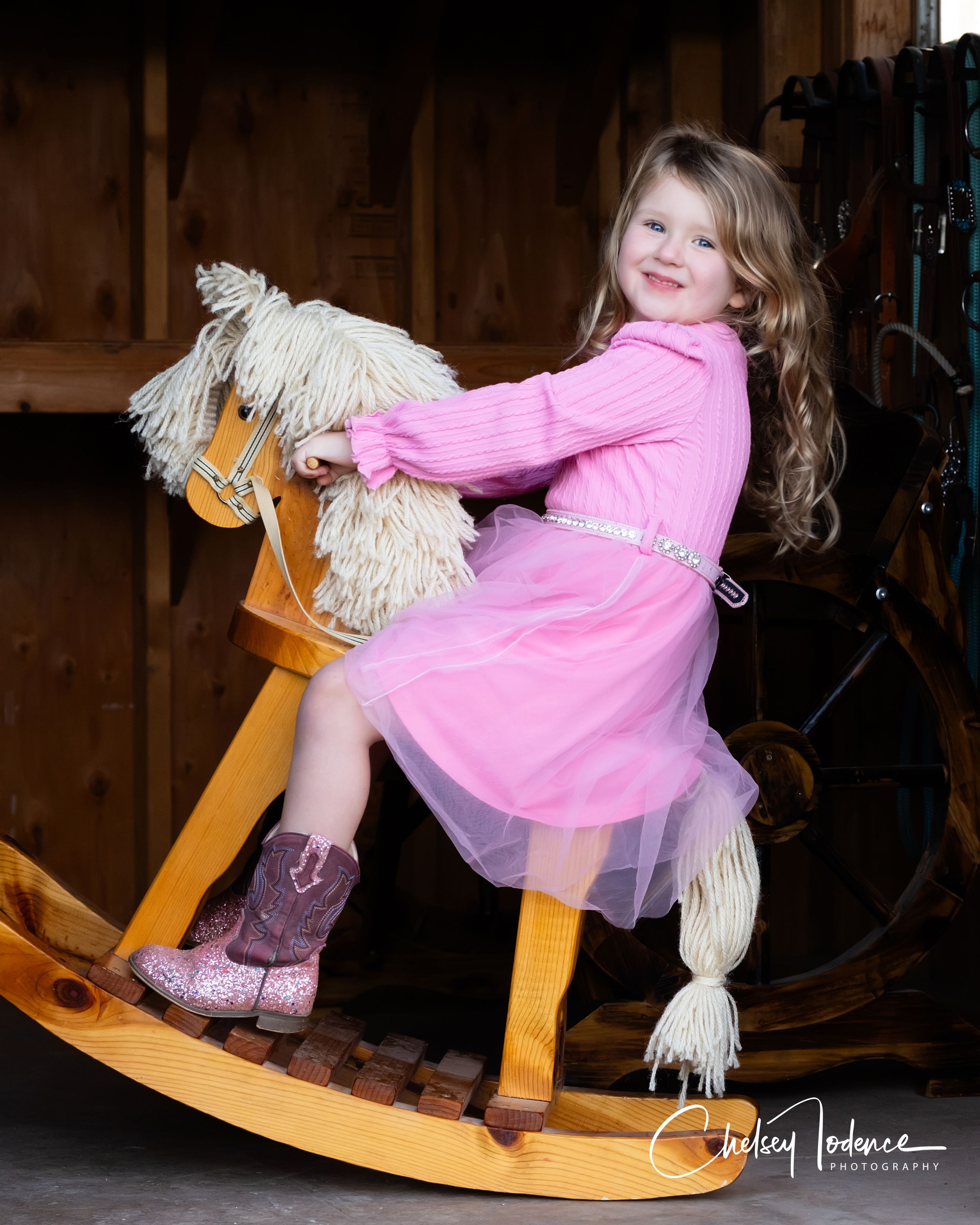 Petaluma CA milestone portrait of smiling toddler girl in pink dress riding a wooden rocking horse inside a rustic barn studio