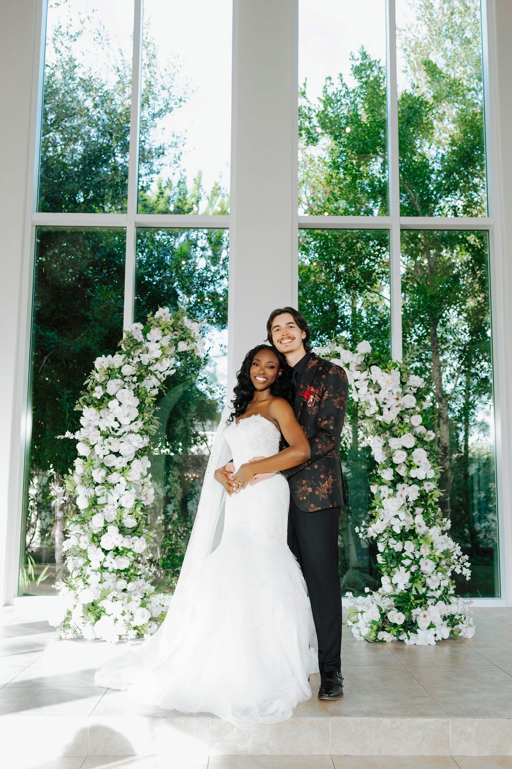 A romantic wedding photoshoot in a bright white room with tall windows and floral decorations.
