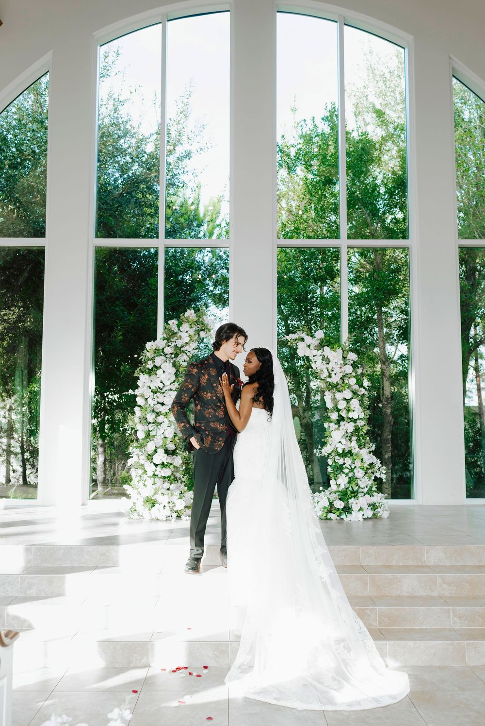 A romantic wedding moment captured in a bright white colonnade with large windows and floral arrangements.