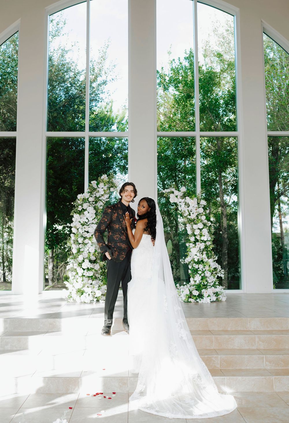 A romantic wedding portrait featuring a couple embracing by a grand window adorned with white floral arrangements.