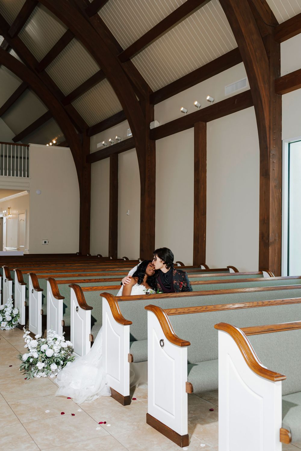 Interior of a modern church chapel with white pews, exposed wooden beams, and floral decorations along the aisle.