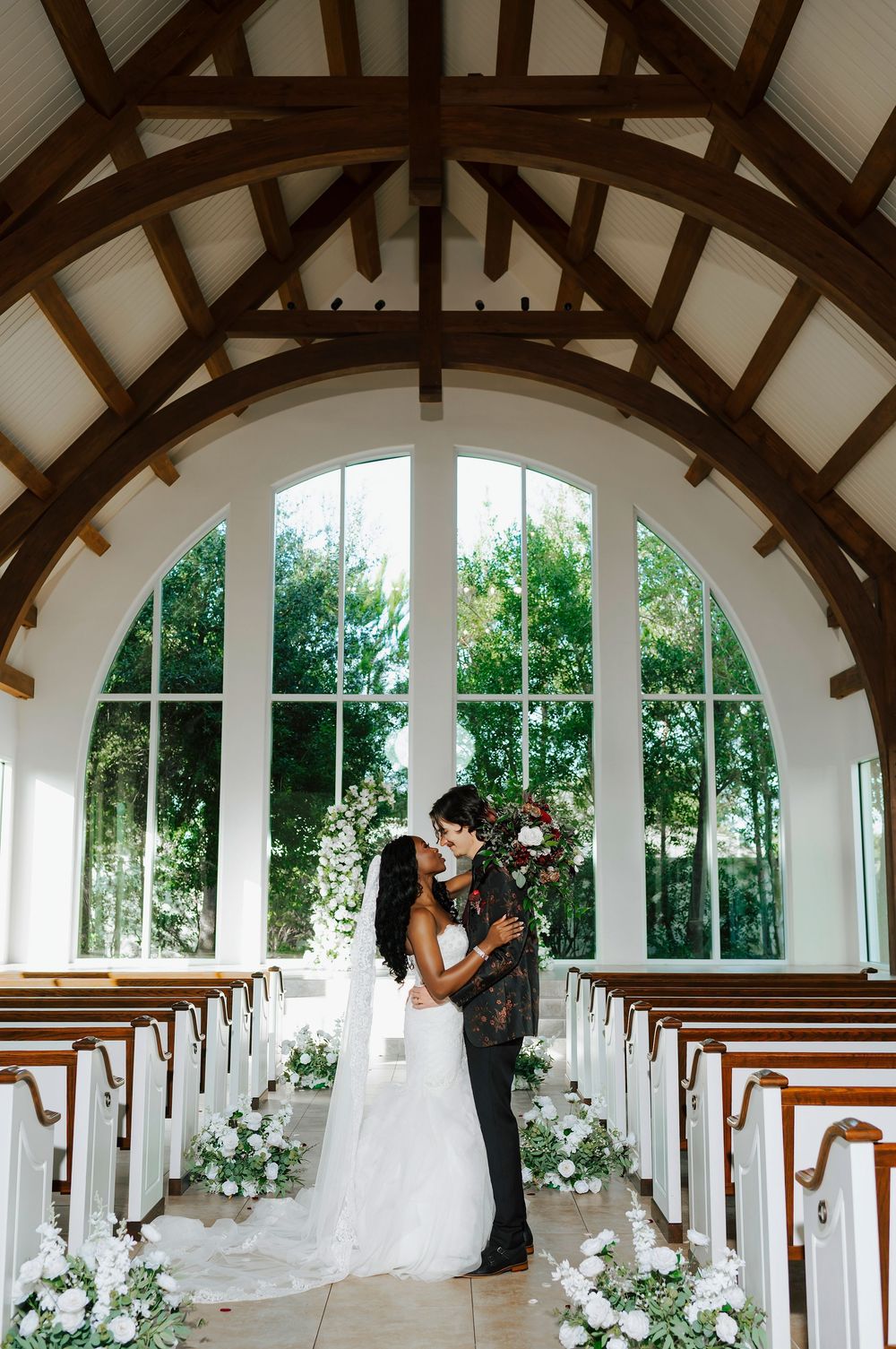 A wedding ceremony venue with wooden beams, white arches, and church pews decorated with elegant floral arrangements.