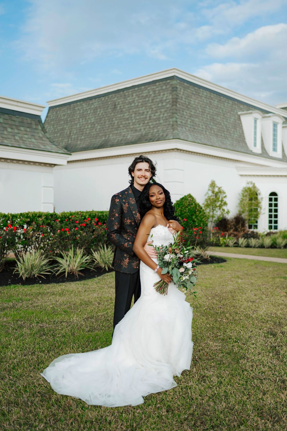 A couple in wedding attire poses together on a grassy lawn with flowering bushes and a white building in the background.