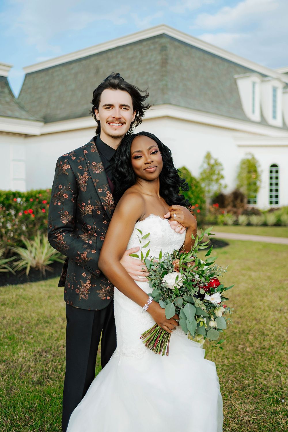 A romantic wedding couple embraces outdoors on a sunny day with a white building in the background.