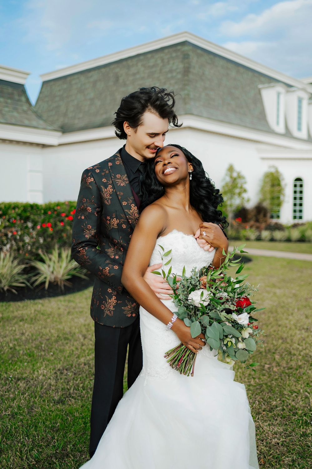 A romantic wedding couple embraces on a lawn with a white church building in the background.