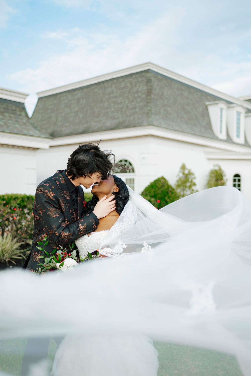 A romantic wedding moment captured through delicate veil movement outside an elegant white building on a sunny day.