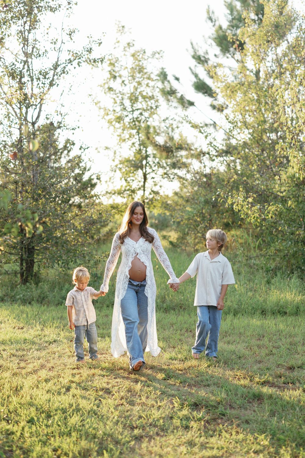 Pregnant woman in white lace cardigan and jeans walks outdoors holding hands with two young boys in a sunny green field.