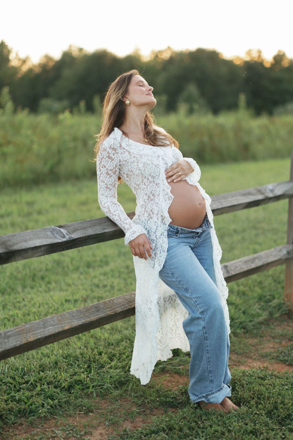 Pregnant woman in white lace top and jeans leaning on wooden fence outdoors, cradling baby bump at golden hour.