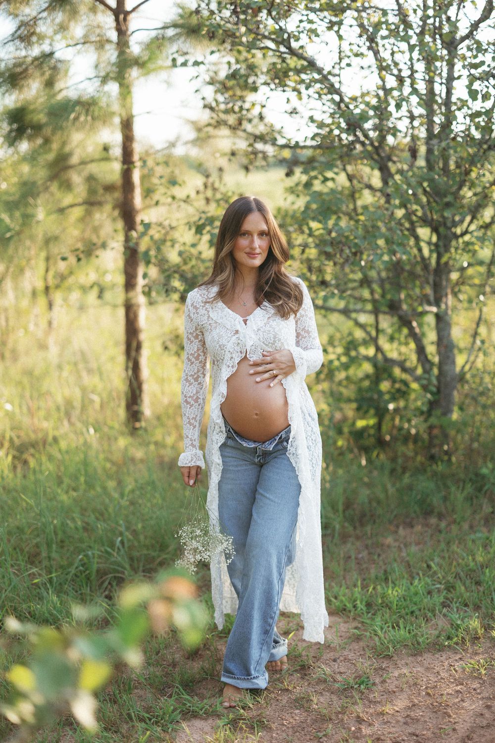 Pregnant woman in white lace cardigan and jeans holds baby bump while walking on a nature path.