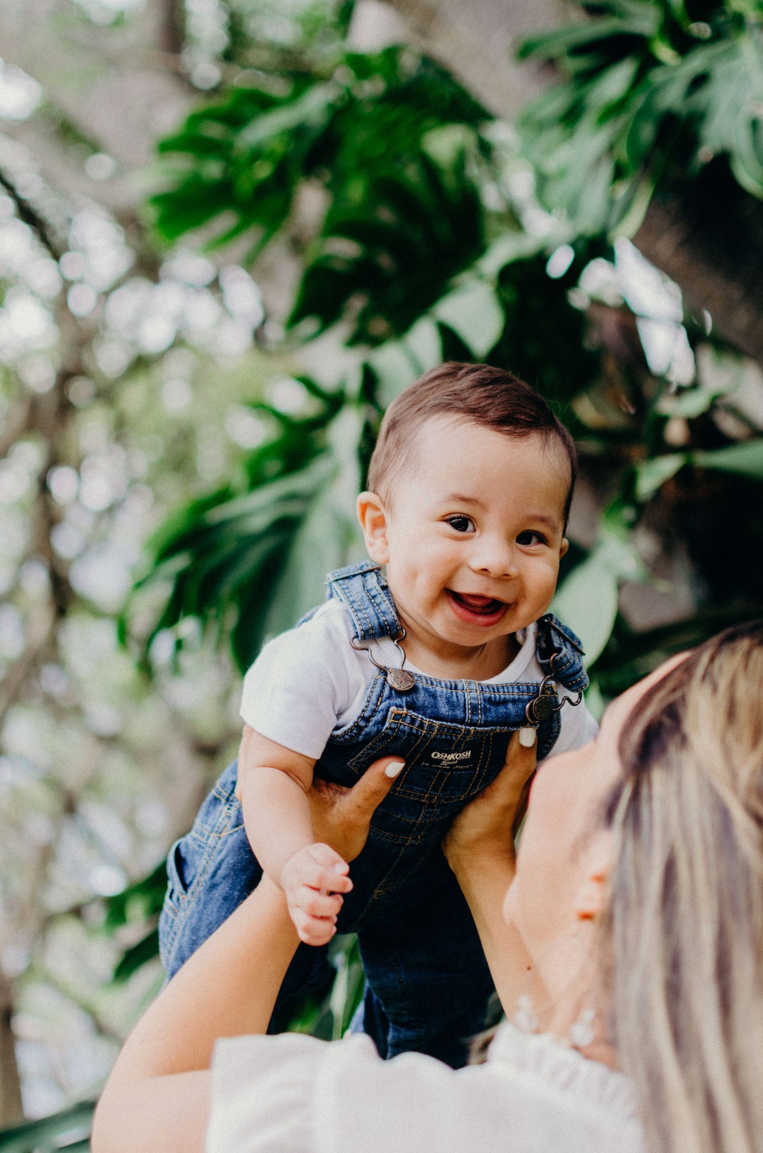 A baby wearing denim overalls smiles brightly while being held up against tropical foliage.