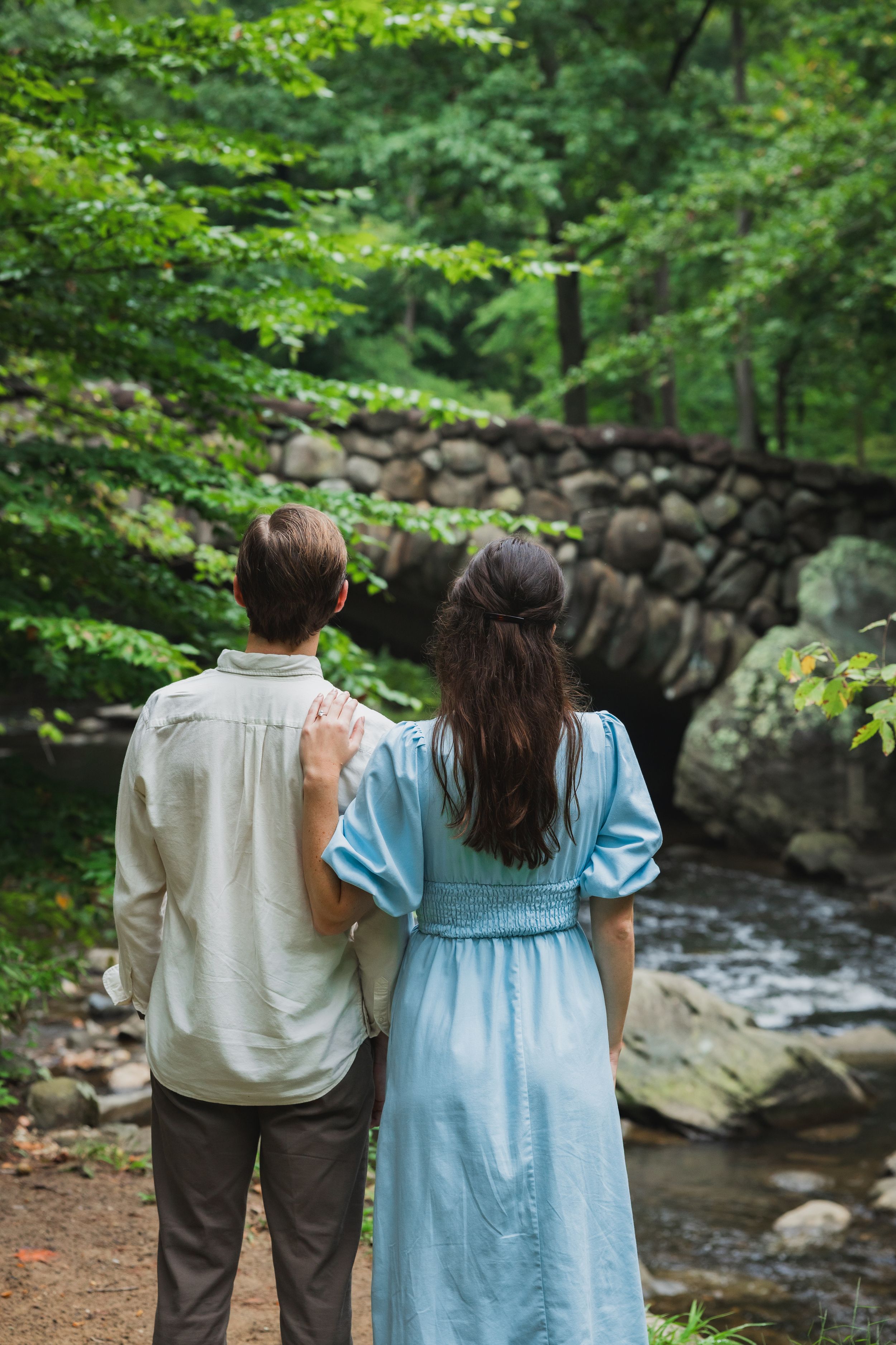 A couple in light blue dress and white shirt stand together overlooking the scenic boulder bridge and stream in a forest setting.