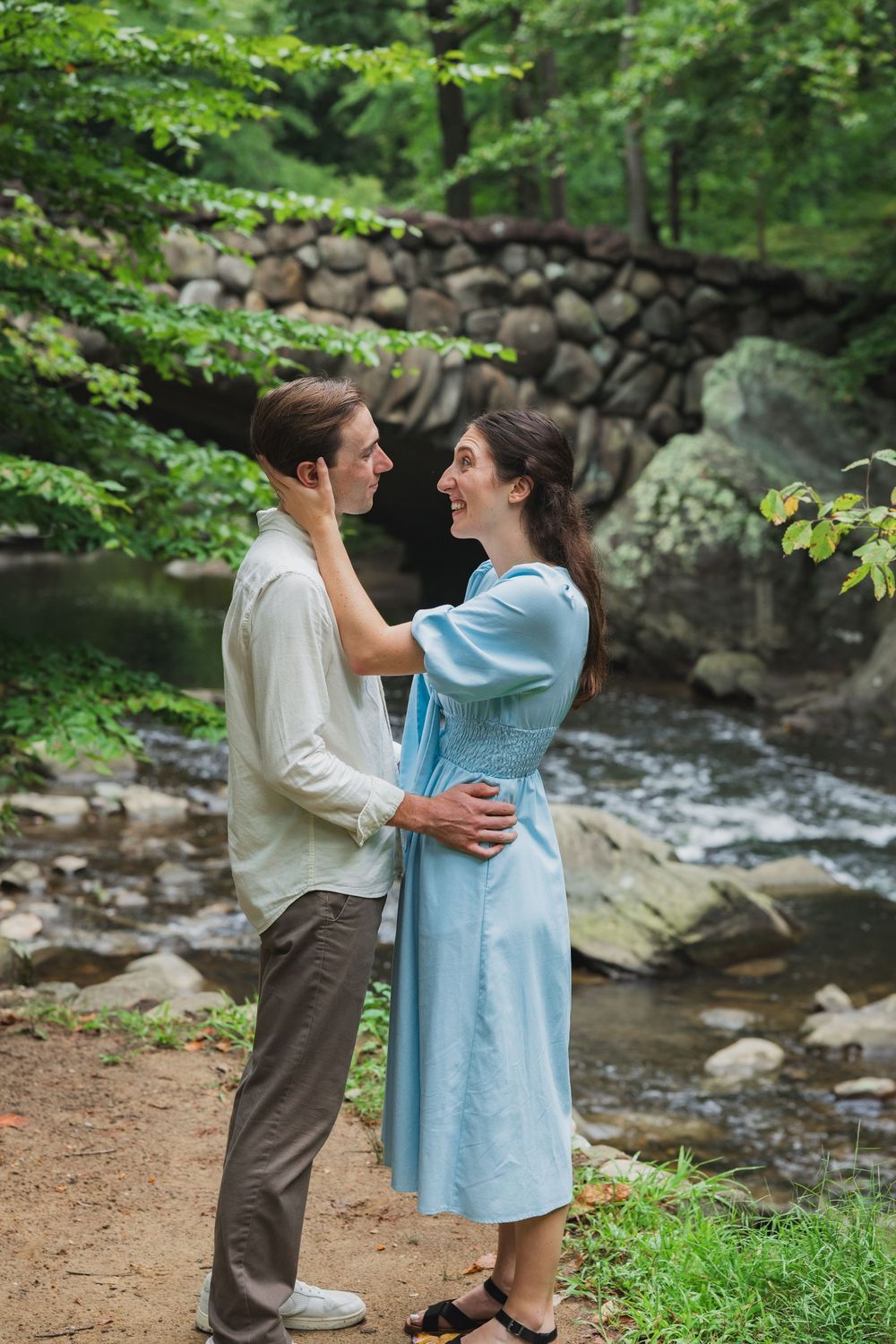 A couple embraces by a scenic forest stream with stone walls and lush greenery in the background at boulder bridge
