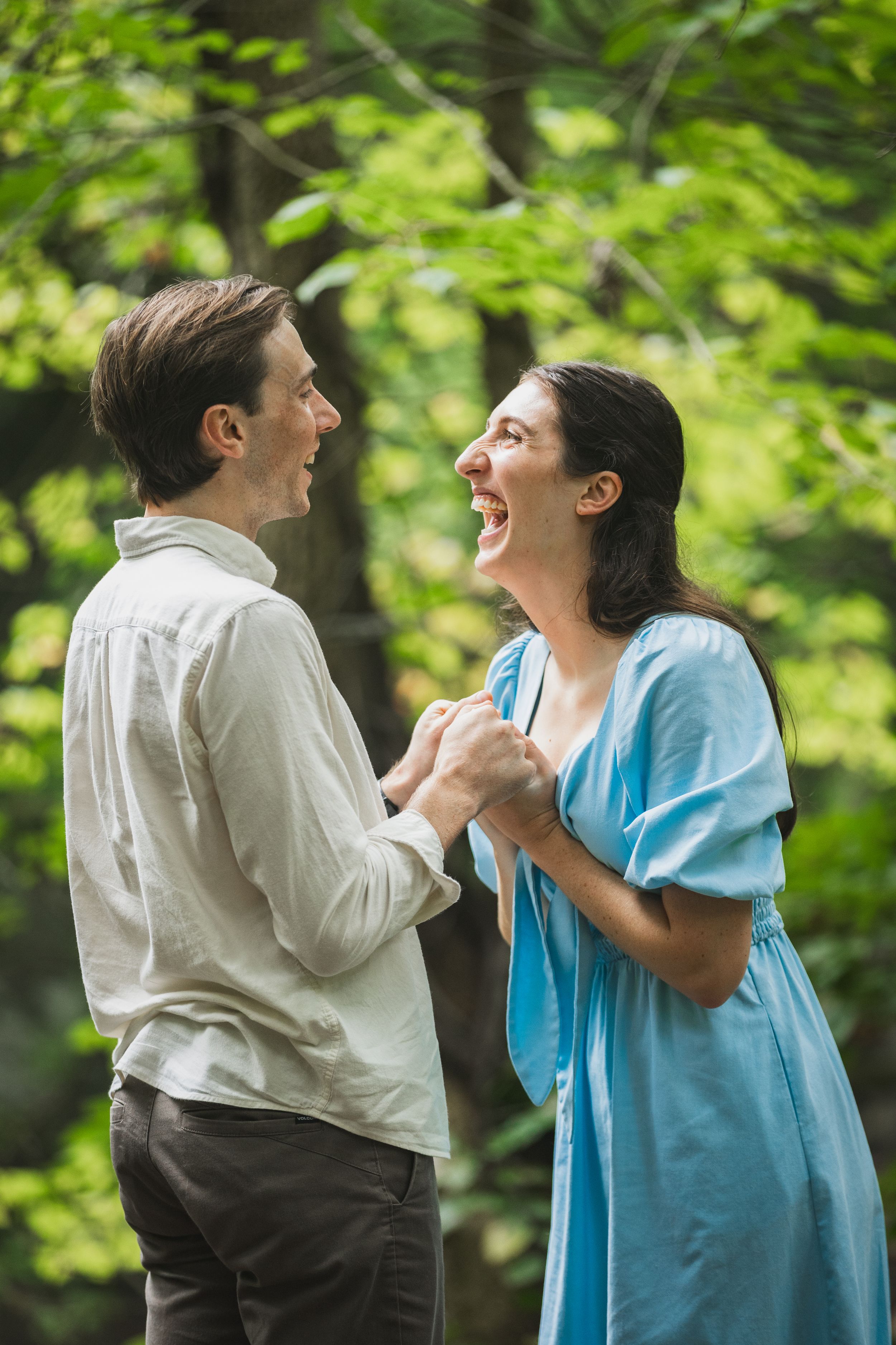 A couple in white and blue clothing share a joyful moment laughing together outdoors against green foliage.