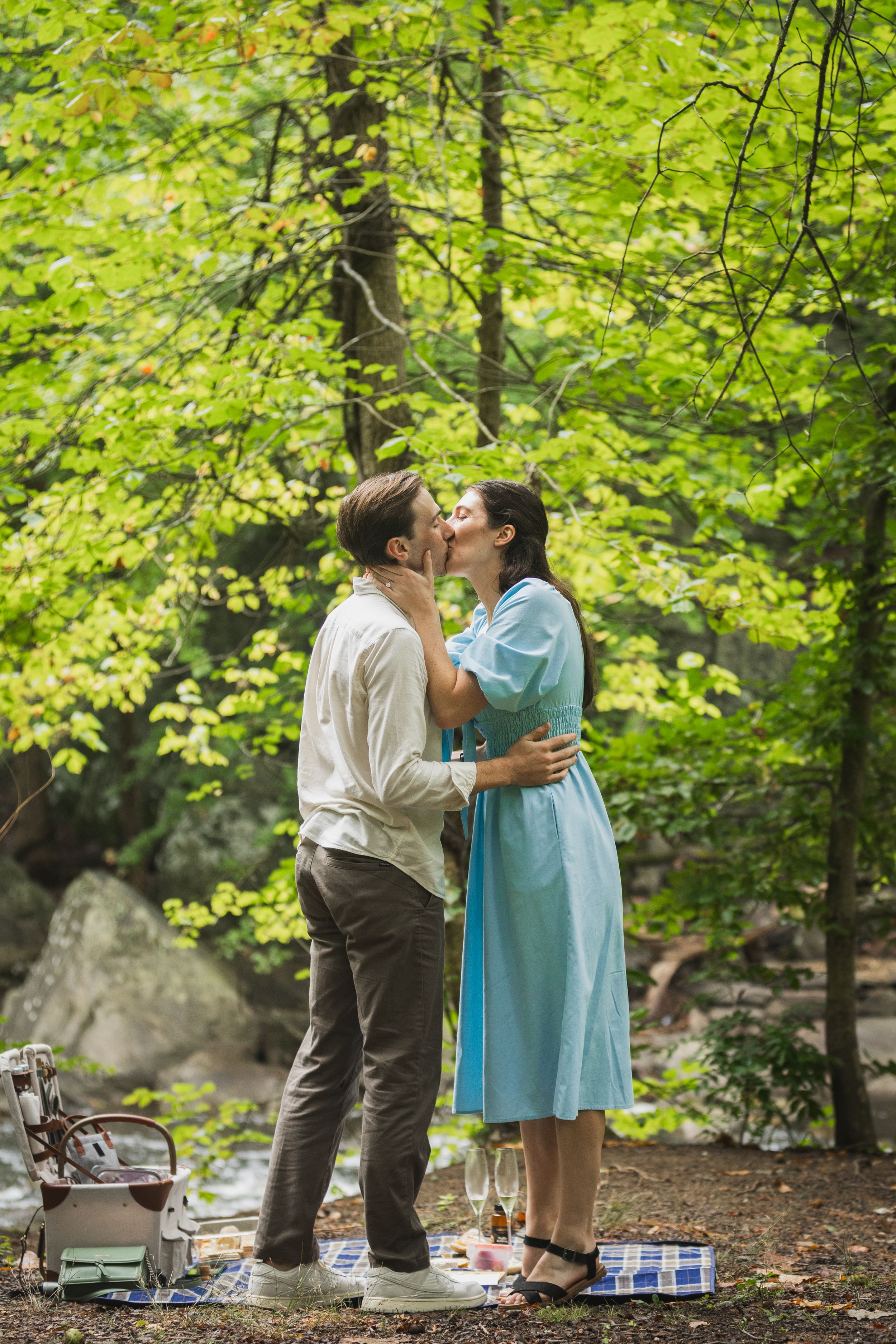 A romantic picnic scene in a lush green forest with champagne and gifts on the ground.