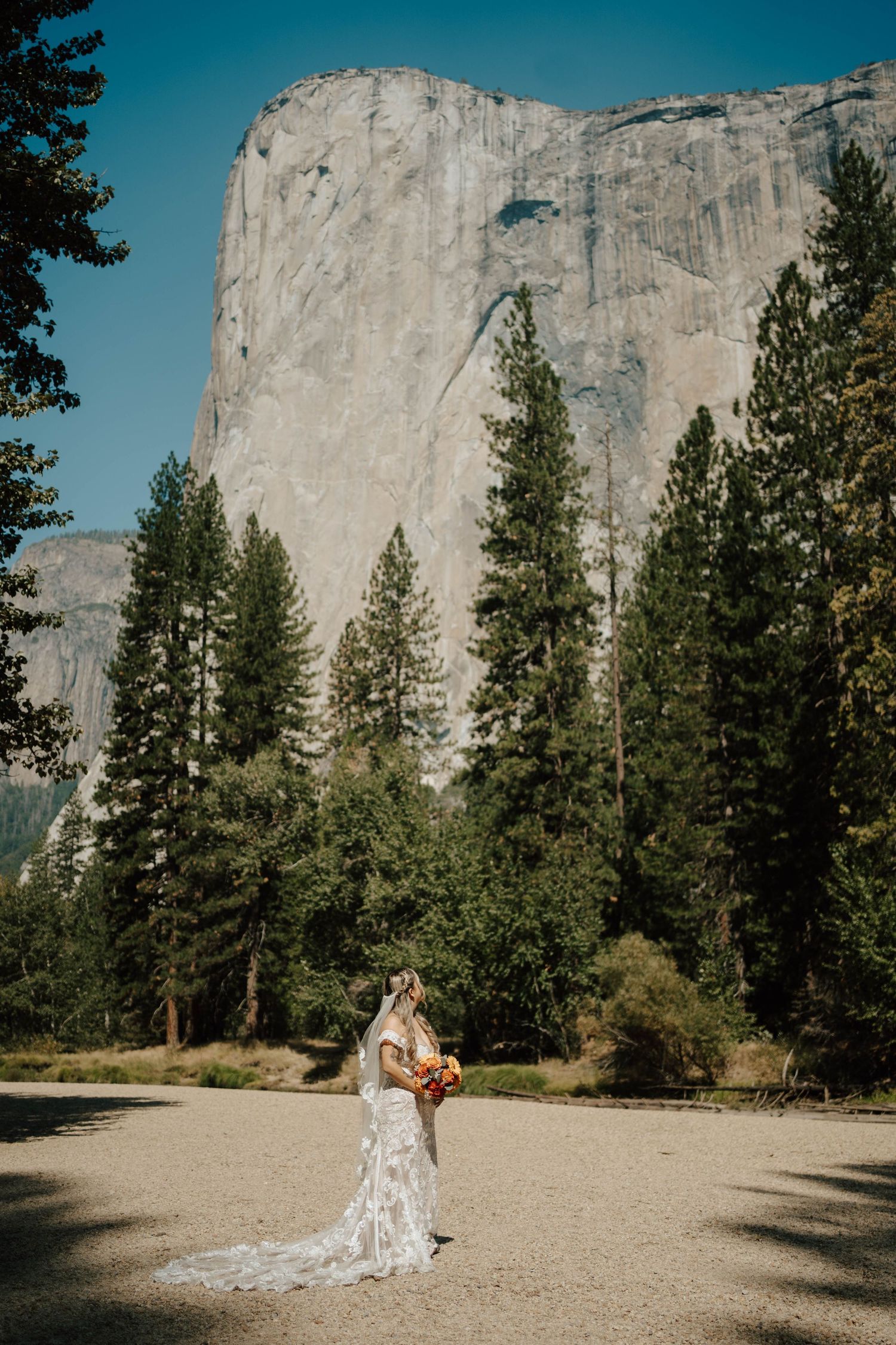 Bride in lace gown with long veil stands before El Capitan in Yosemite National Park.