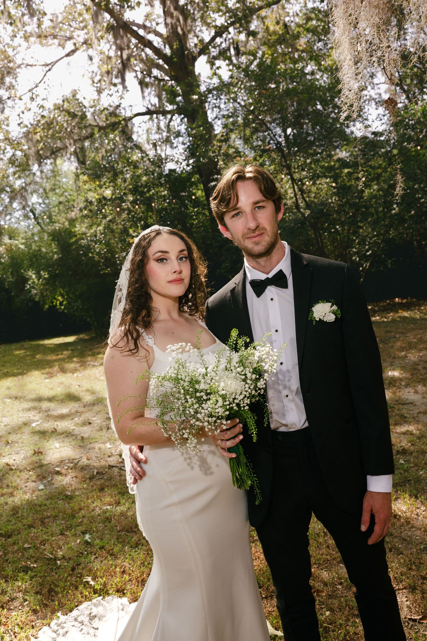 A couple in formal wedding attire poses outdoors beneath Spanish moss-draped trees on their special day at The Elms of Coosada.