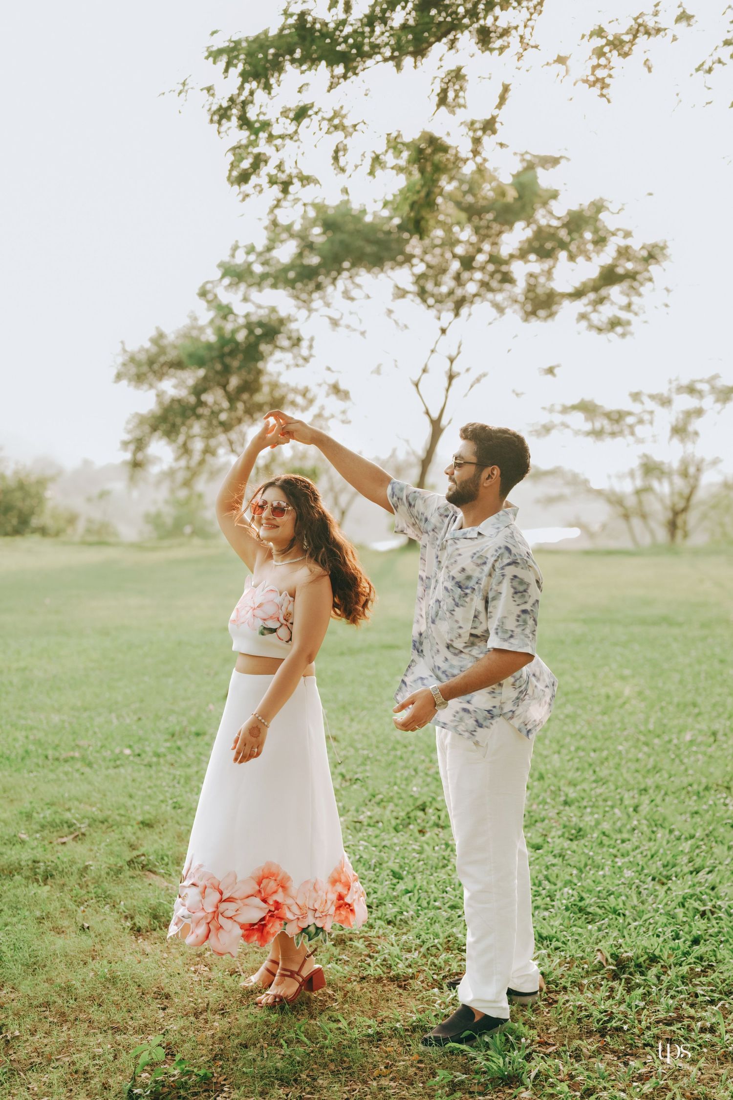 A couple dances together in a grassy field during sunset, wearing white and floral outfits.