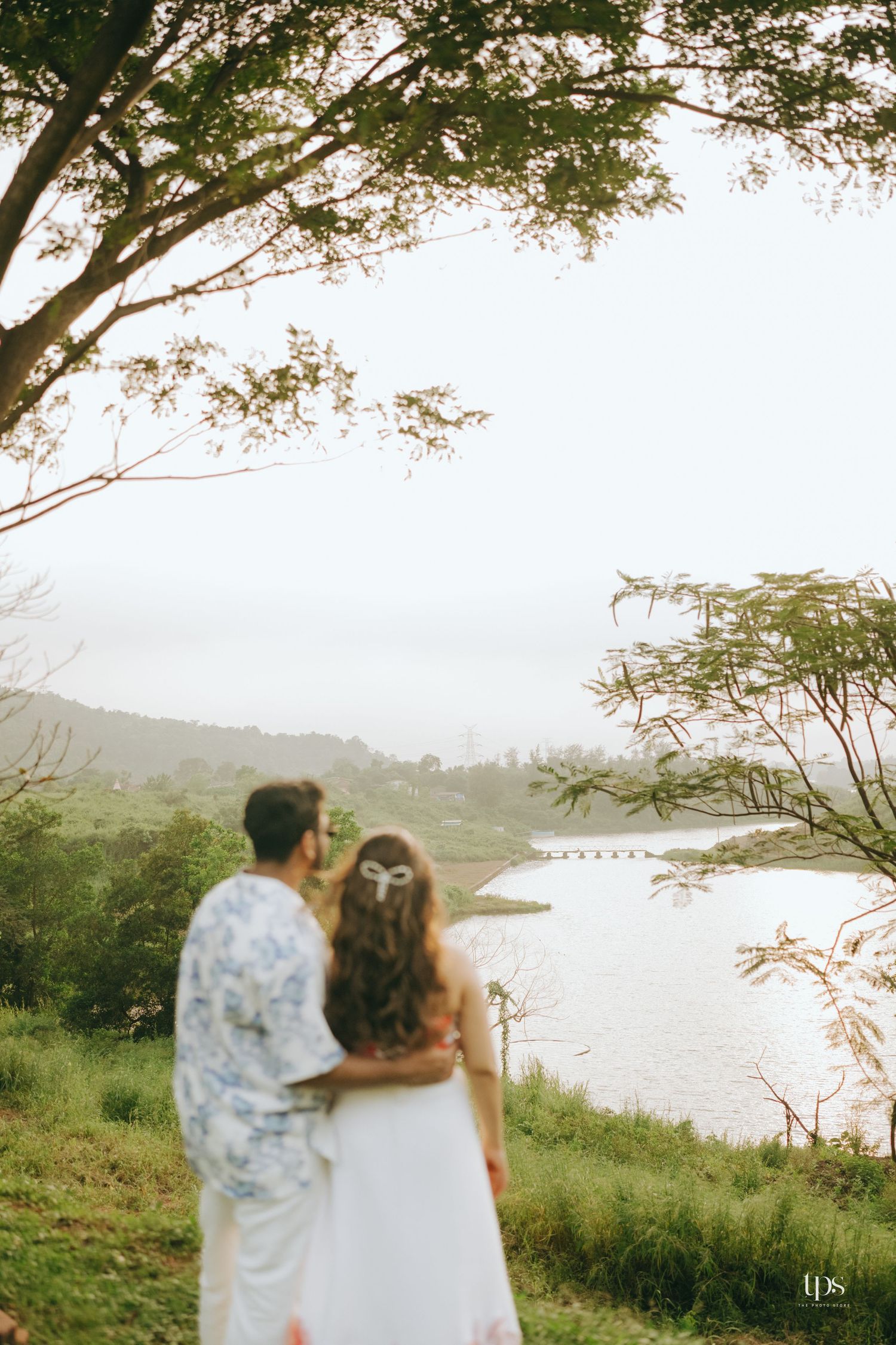 A couple stands together overlooking a scenic coastal view with trees framing the shot.
