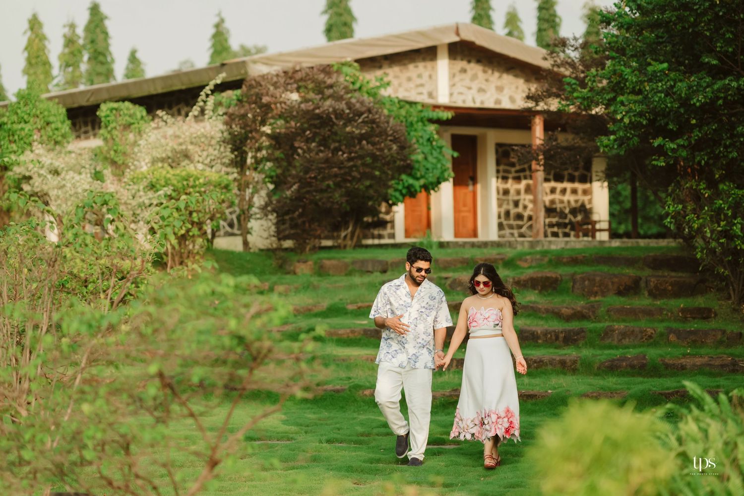 A couple strolls through a garden path with a rustic stone building in the background.