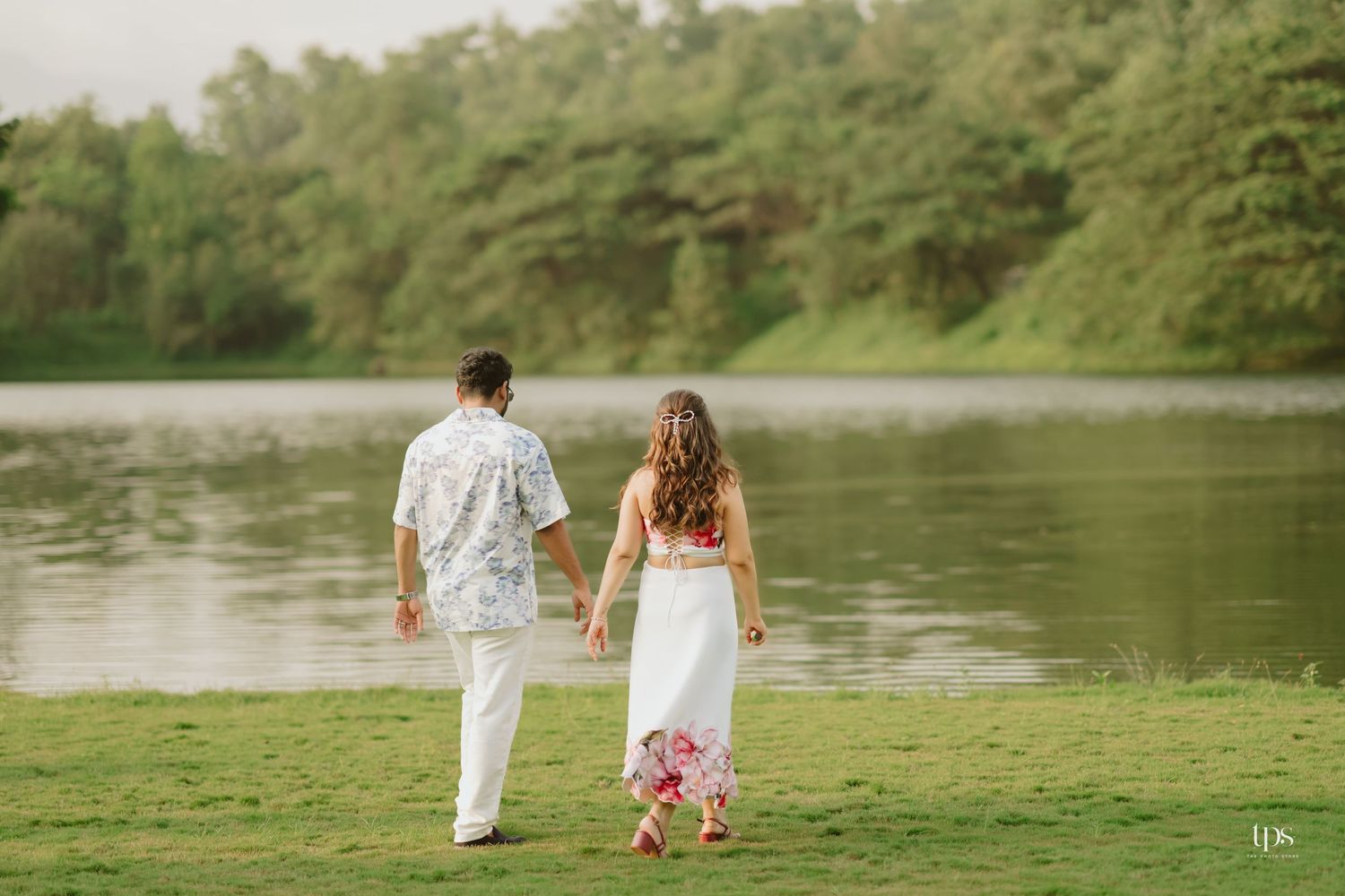 A couple walks along a lakeside path surrounded by lush greenery and trees.